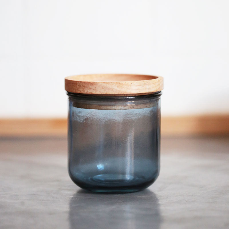 Ein durchscheinend blaues Glasgefäß mit einem runden Holzdeckel steht auf einer glatten grauen Oberfläche vor einem sanft verschwommenen Hintergrund.