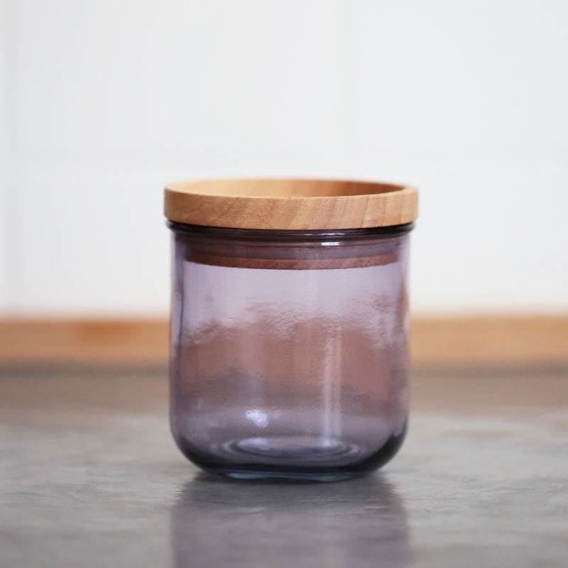 Ein transparentes lila Glas mit einem glatten Holzdeckel steht auf einer grauen Fläche vor einem unscharfen weißen Hintergrund.