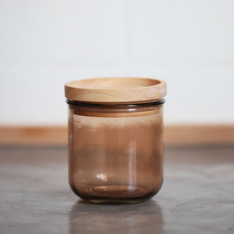 Ein braunes Glasgefäß mit einem runden Holzdeckel steht auf einer grauen Fläche vor einem weißen Hintergrund.