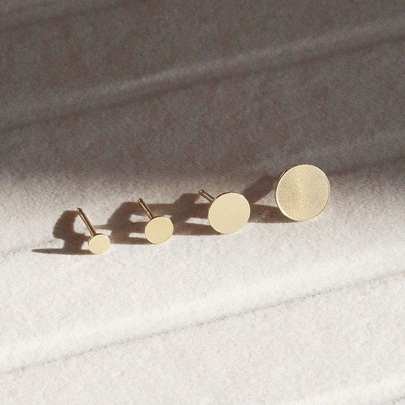 Vier runde goldene Ohrstecker mit zunehmender Größe sind in einer Reihe auf einer strukturierten, hellen Oberfläche mit weichen Schatten angeordnet.