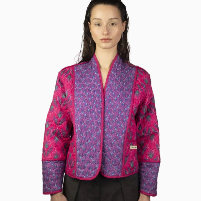 Eine Frau mit nassem, nach hinten gegeltem Haar trägt eine leuchtende Steppjacke mit einem geometrischen Muster in Rosa und Lila. Sie steht vor einem schlichten, hellen Hintergrund und schaut direkt in die Kamera.