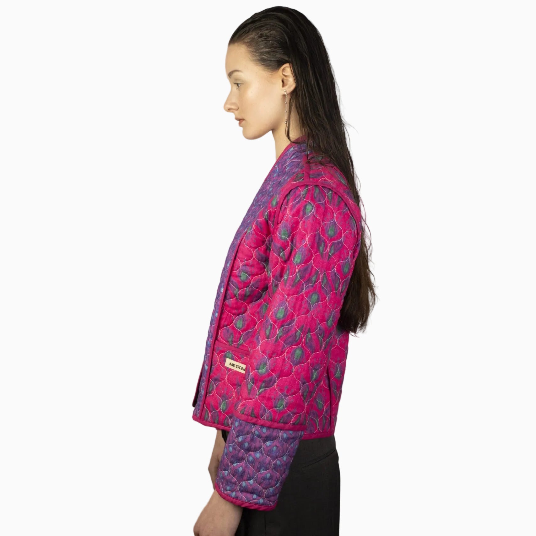 Eine Frau mit langen, nass aussehenden braunen Haaren steht im Profil und trägt eine leuchtend rosa und violette Steppjacke mit einem Blattmuster und eine dunkle Hose vor einem schlichten hellen Hintergrund.