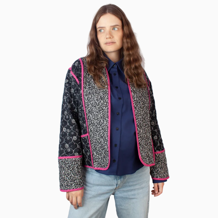 Eine Person mit langen braunen Haaren trägt ein dunkelblaues Button-up-Hemd, hellblaue Jeans und eine gemusterte schwarz-weiße Jacke mit leuchtend rosafarbenem Rand. Sie steht vor einem hellen Hintergrund und schaut leicht zur Seite.