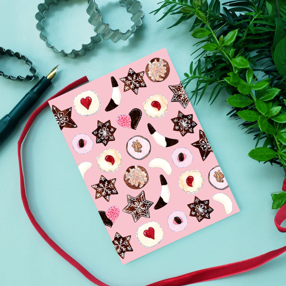 Eine Grußkarte mit verschiedenen illustrierten Keksen auf rosafarbenem Hintergrund liegt auf einer hellen Fläche, daneben ein rotes Band, ein Stift, Ausstechformen aus Metall und grüne Pflanzen.