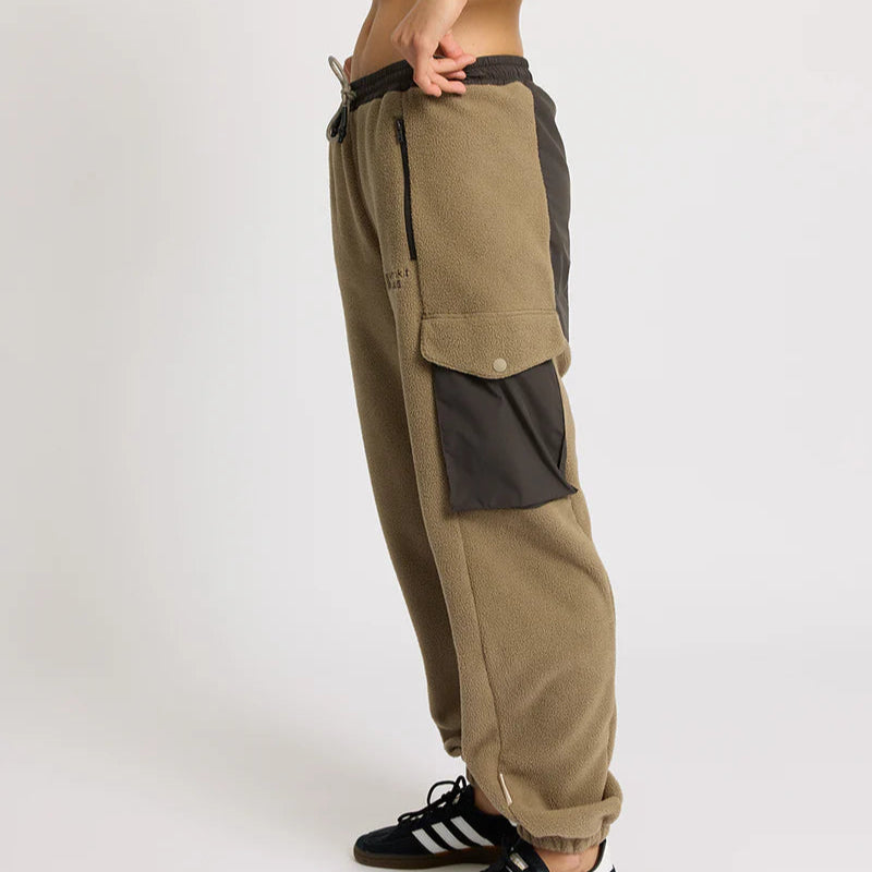 Eine Person, die einen schwarzen Sport-BH, eine hellbraune Fleece-Cargohose mit großen Seitentaschen und schwarze Adidas-Turnschuhe trägt, steht seitlich vor einem schlichten weißen Hintergrund.
