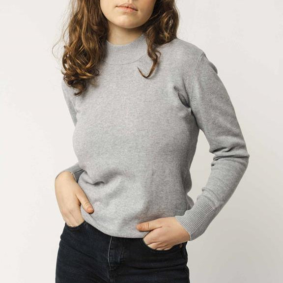 Eine Person mit gewelltem braunem Haar trägt einen hellgrauen langärmeligen Pullover und eine schwarze Hose und steht mit den Händen in den Taschen vor einem schlichten hellen Hintergrund. Ihr Gesicht ist teilweise außerhalb des Rahmens.