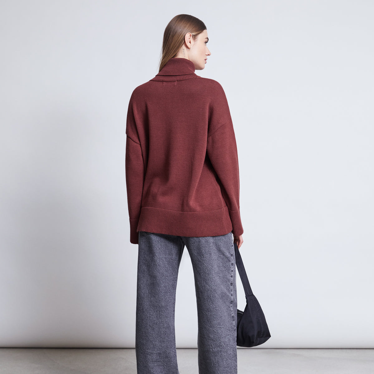 Eine Frau steht mit dem Gesicht zur Kamera, trägt einen lockeren kastanienbraunen Rollkragenpullover und eine graue Hose mit weitem Bein und hält eine schwarze Tasche vor einem schlichten hellen Hintergrund.
