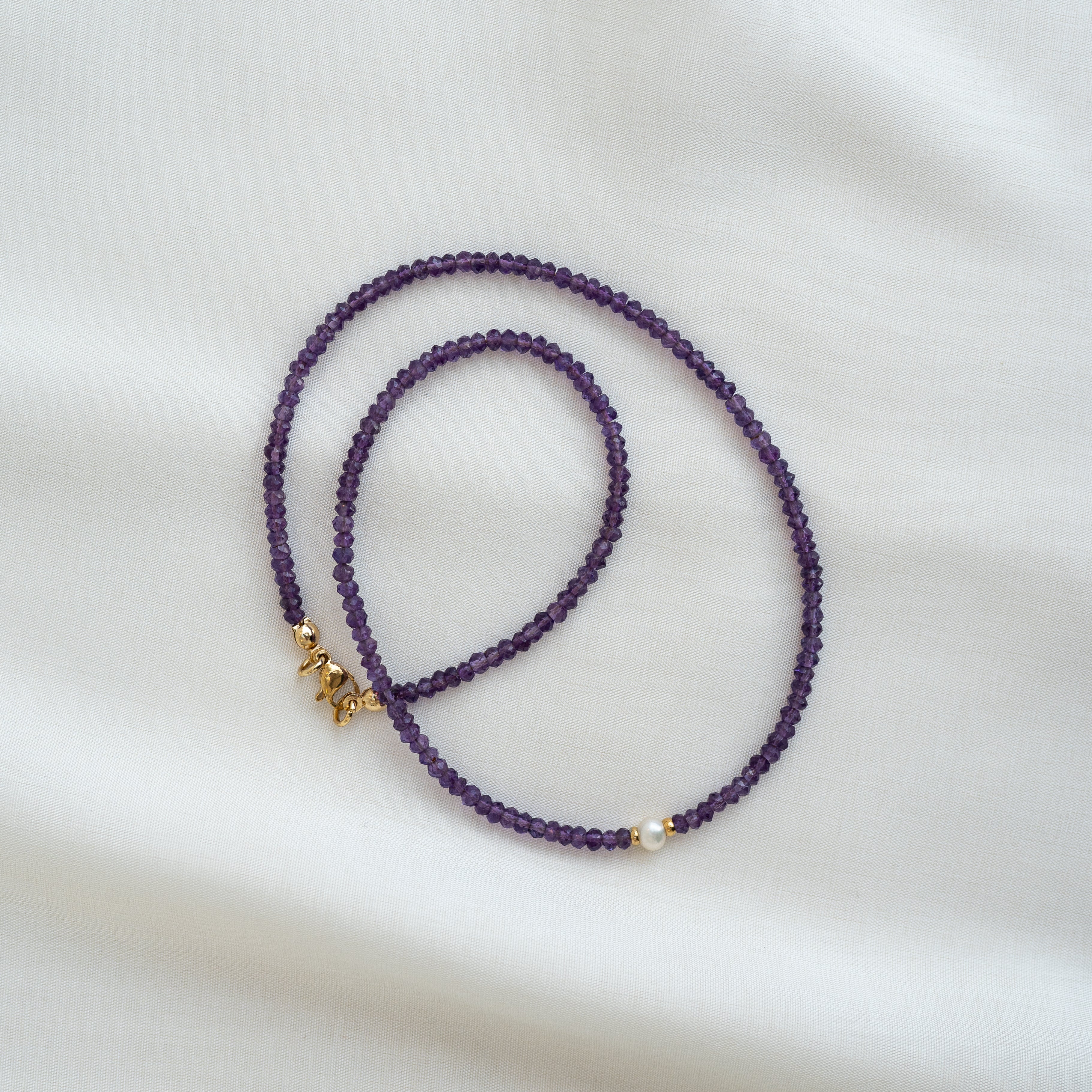 Eine zarte Halskette aus kleinen lila Perlen ist in einer Schleife auf einem weißen Stoffhintergrund angeordnet. Sie enthält eine einzelne Perle und kleine goldene Buchstaben, die Liebe bedeuten.