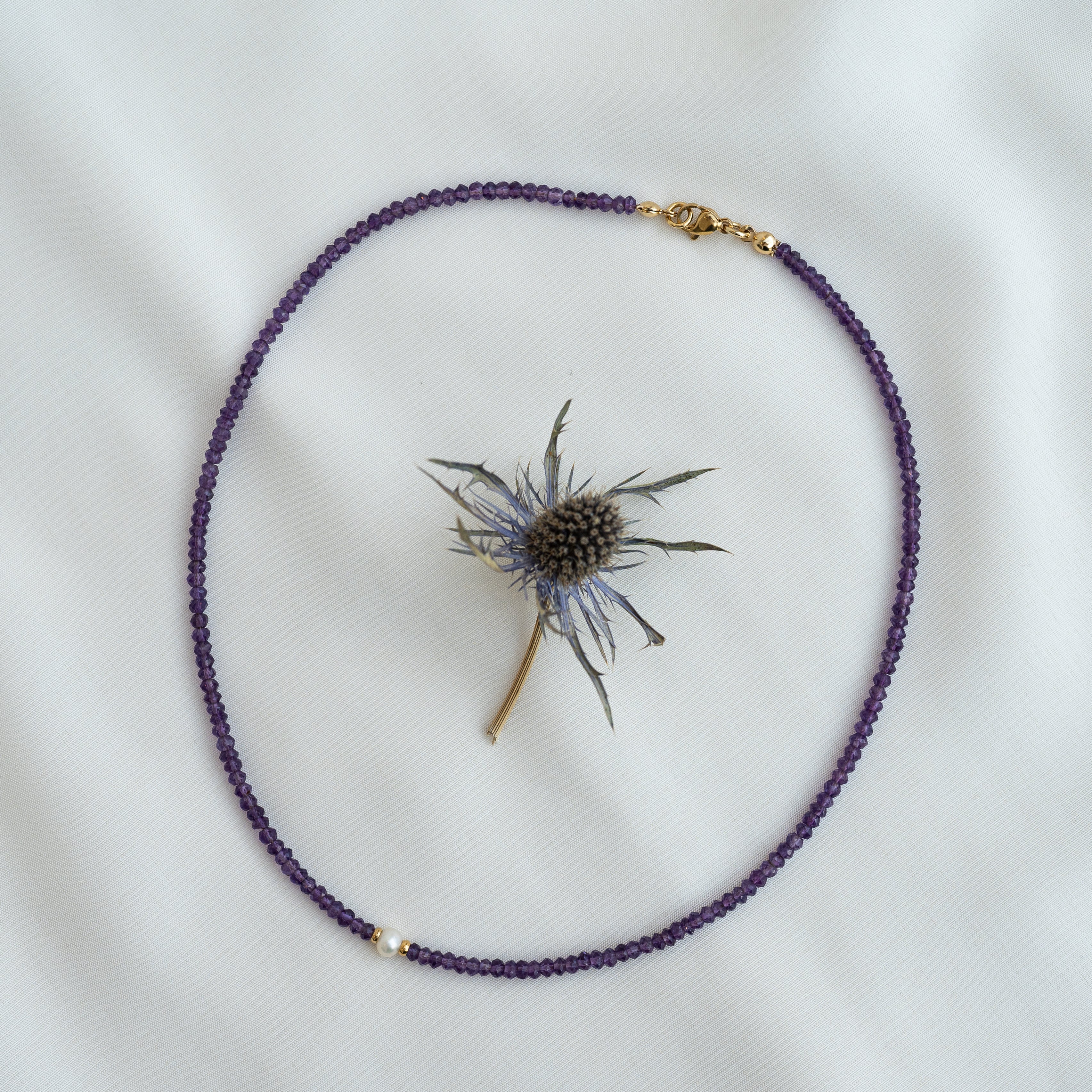 Eine zarte lilafarbene Perlenkette mit einem goldenen Verschluss ist kreisförmig um eine kleine getrocknete Distelblüte angeordnet, alles auf einem weichen weißen Stoffhintergrund.