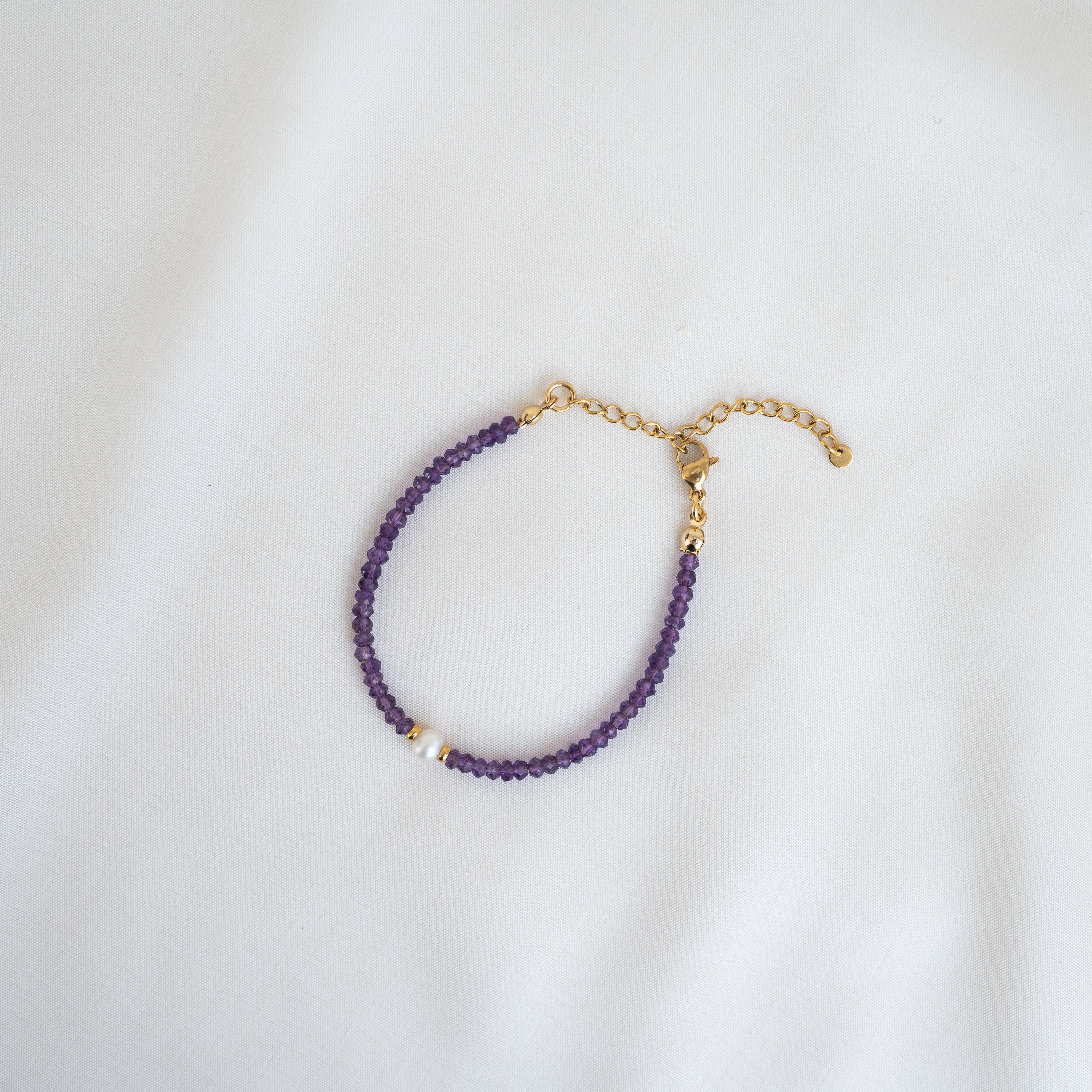 Ein zartes Armband mit kleinen lilafarbenen Perlen, einer einzelnen weißen Perle und einem goldenen Kettenverschluss auf einem weißen Stoffhintergrund.