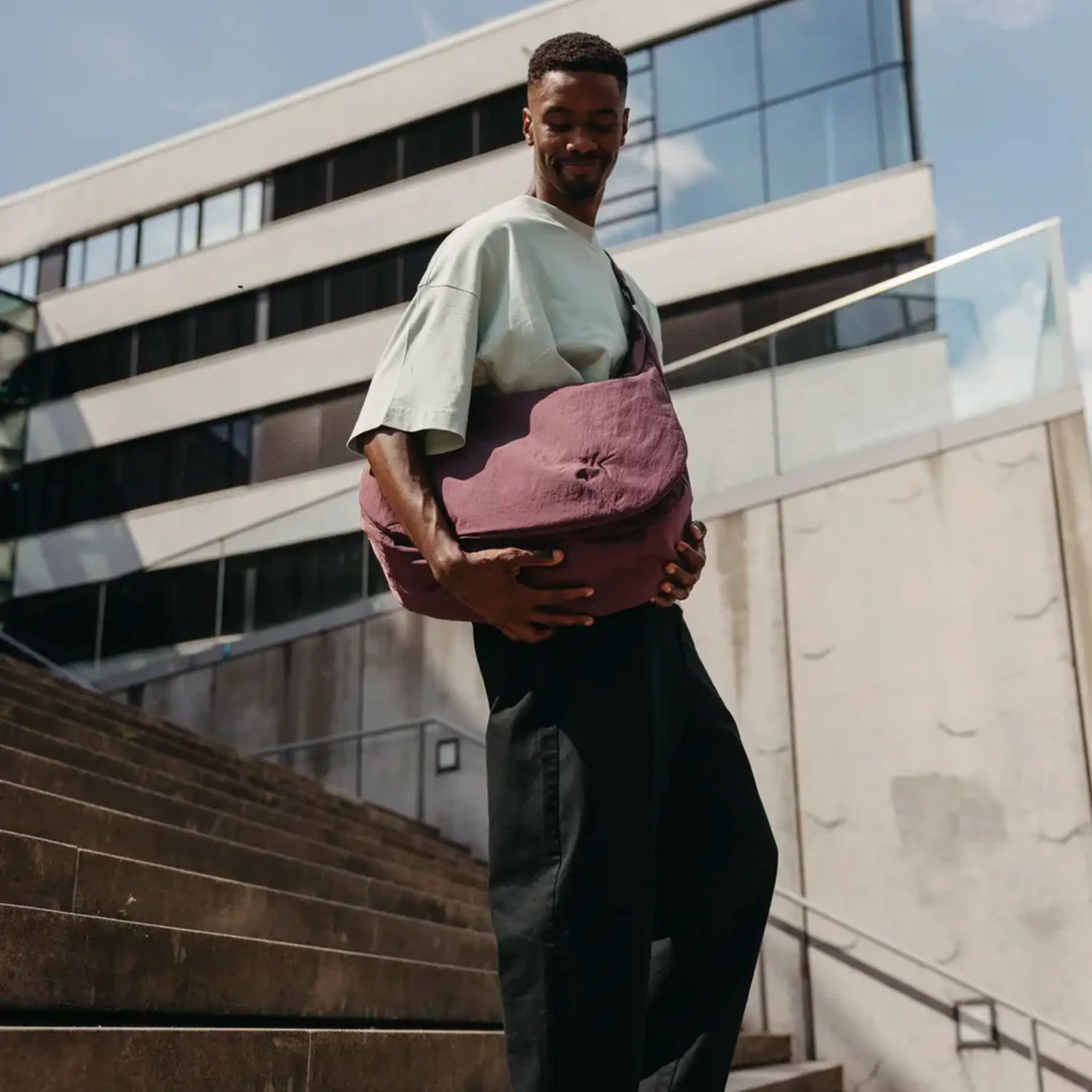 Ein Mann, der ein lockeres helles T-Shirt und eine dunkle Hose trägt, steht auf einer Außentreppe und hält eine große lilafarbene Tasche in der Hand, während im Hintergrund ein modernes Gebäude aus Glas und Beton zu sehen ist.