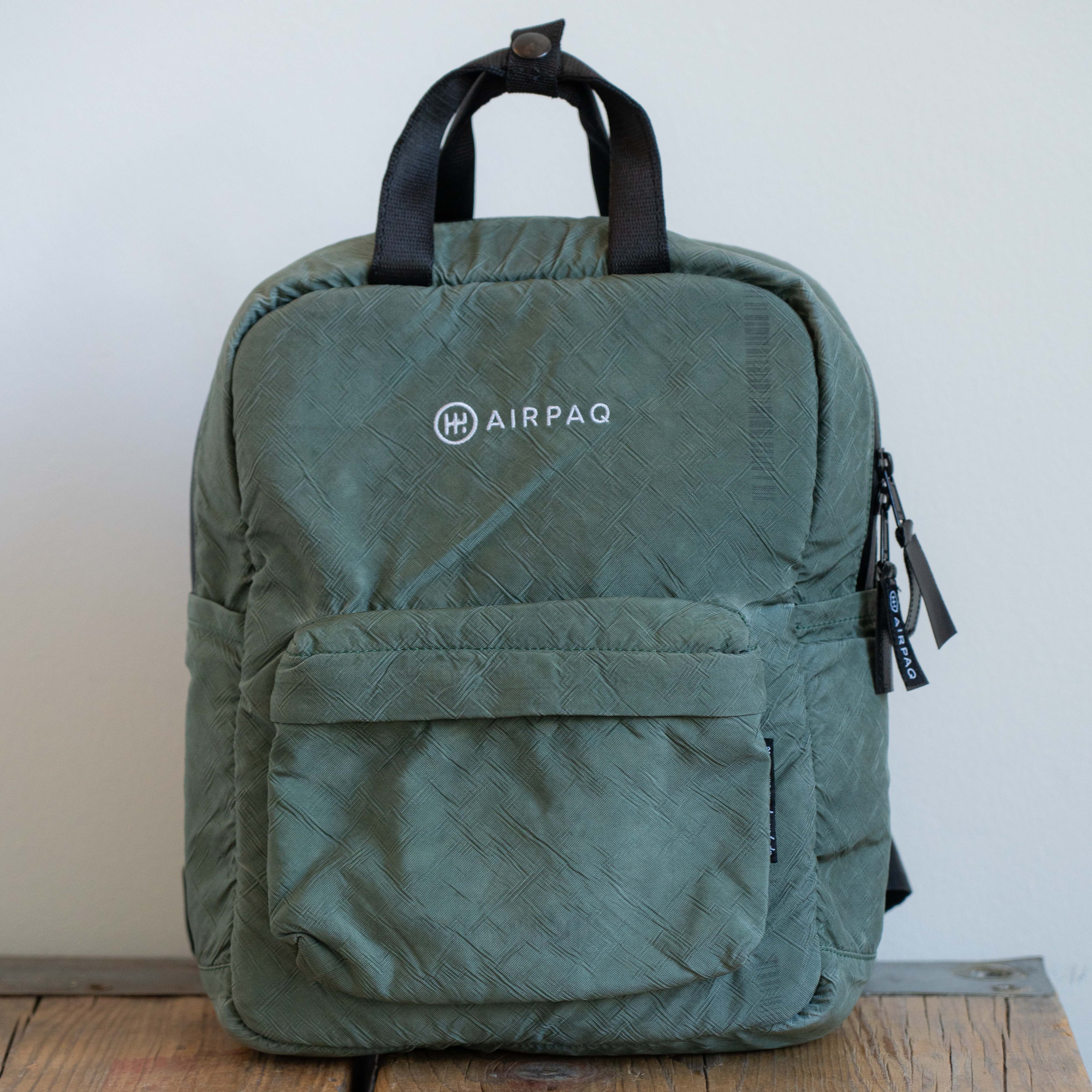 Grüner Rucksack der Marke Airpaq mit schwarzen Trägern, auf einer Holzfläche platziert.