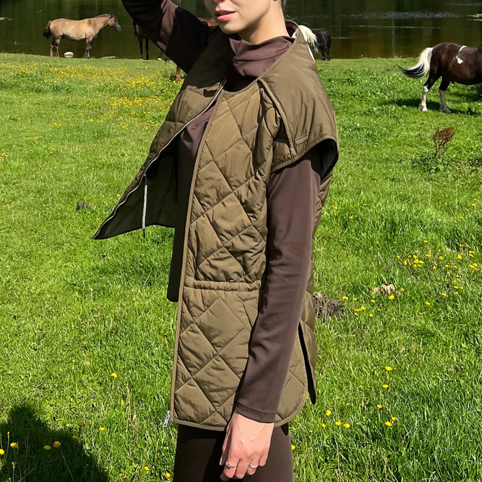 Eine Person in einer gesteppten olivgrünen Weste und einem braunen langärmeligen Oberteil steht auf grünem Gras, während im Hintergrund an einem sonnigen Tag Pferde an einem Teich grasen.