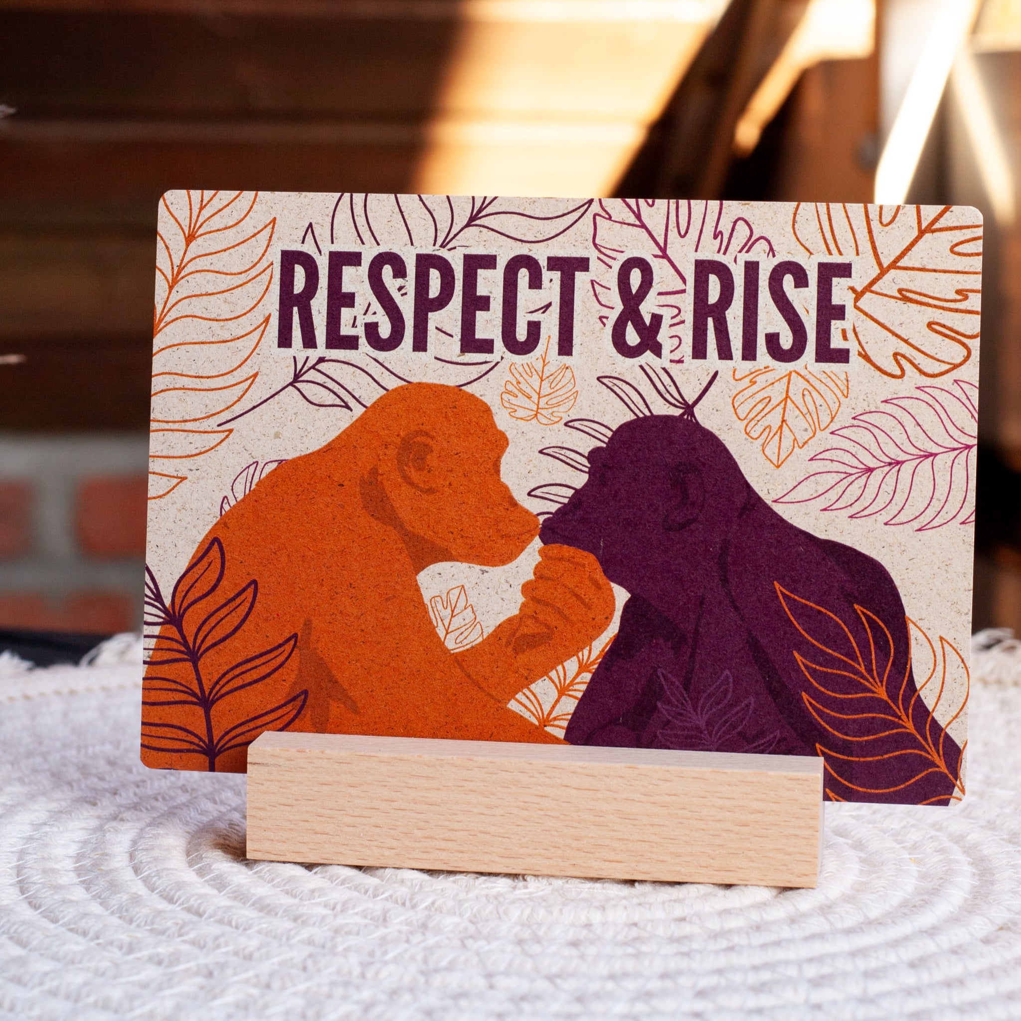 Eine Karte mit Abbildungen von zwei Affen, die sich gegenüberstehen und die Hände halten, einer orange und einer lila, mit dem Text RESPECT & RISE über ihnen, umgeben von Blattmustern, auf einem Holzständer.