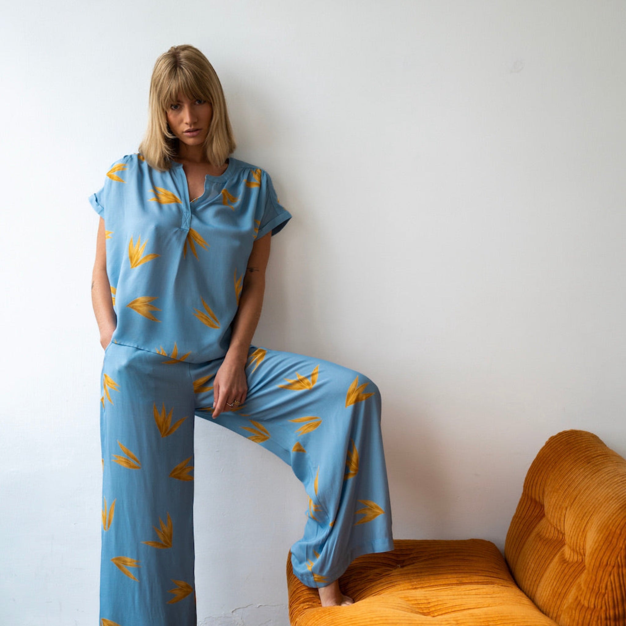 Frau in blauem, bedrucktem Pyjama steht lässig neben einem senffarbenen Sofa vor einer weißen Wand.