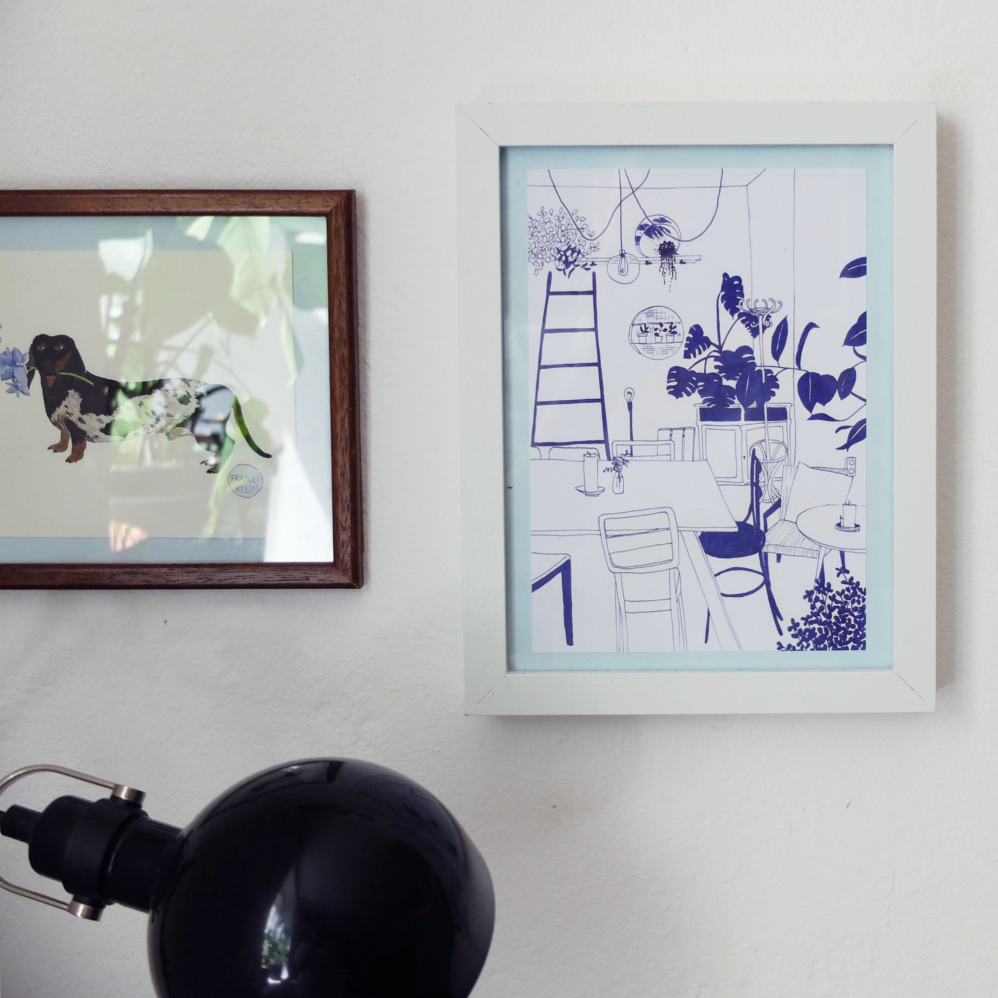 Zwei gerahmte Kunstwerke hängen an einer weißen Wand über einer schwarzen Lampe. Das eine Bild zeigt einen Hund mit Pflanzenmustern, das andere ist eine blaue Strichzeichnung eines Zimmers mit Pflanzen, einer Leiter und Stühlen.