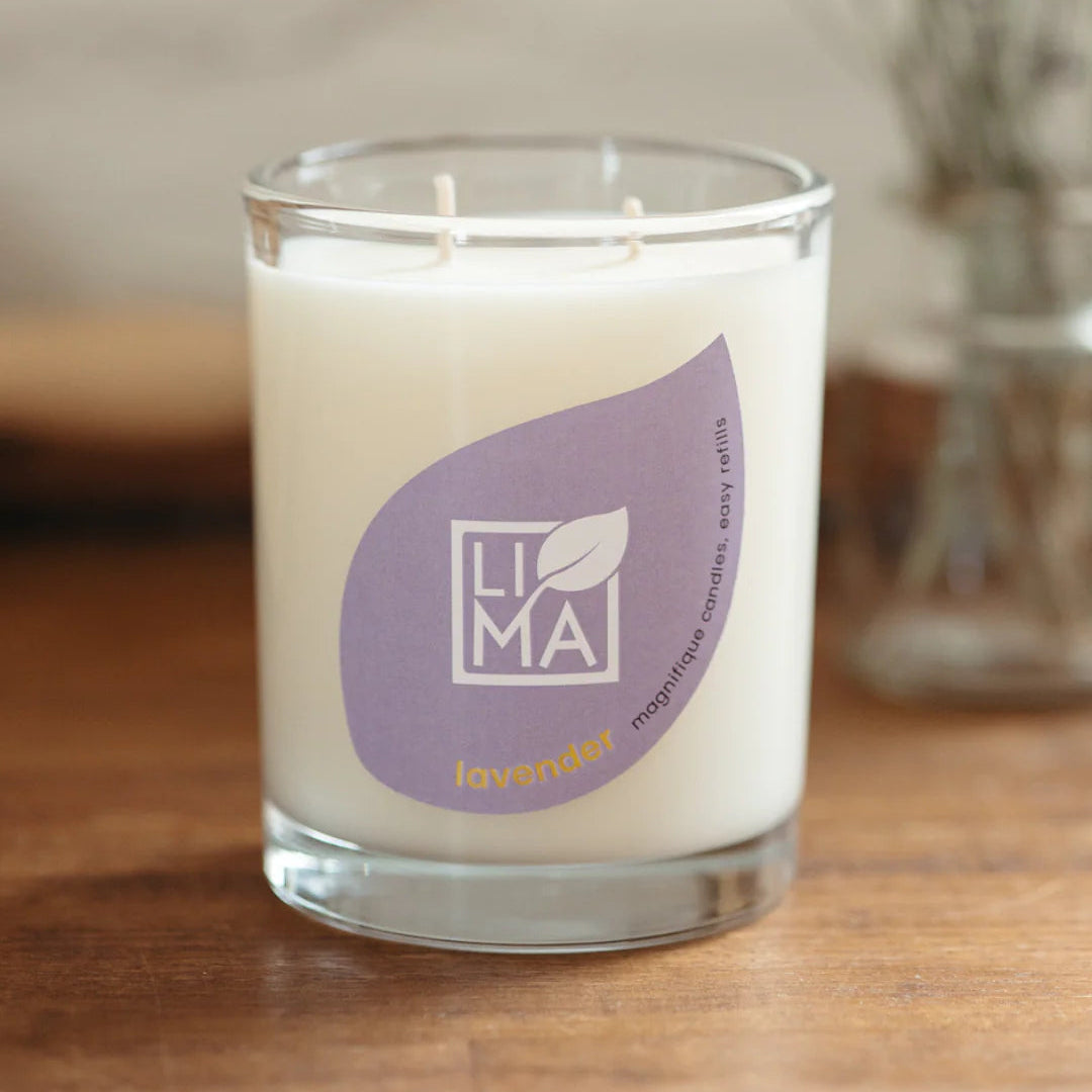 Eine weiße Sojakerze in einem klaren Glasgefäß steht auf einer Holzfläche. Das Etikett der Kerze ist blassviolett und trägt den Markennamen LIMA sowie das Wort Lavendel in Gelb, das auf den Duft hinweist.