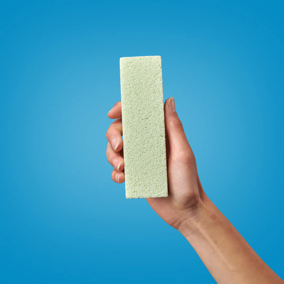 Eine Hand hält einen rechteckigen Bimsstein senkrecht vor einem einfarbig blauen Hintergrund.