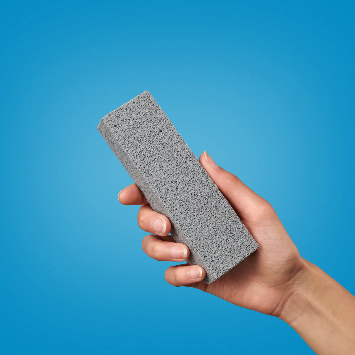 Eine Hand hält einen rechteckigen grauen Bimsstein vor einem einfarbig blauen Hintergrund.
