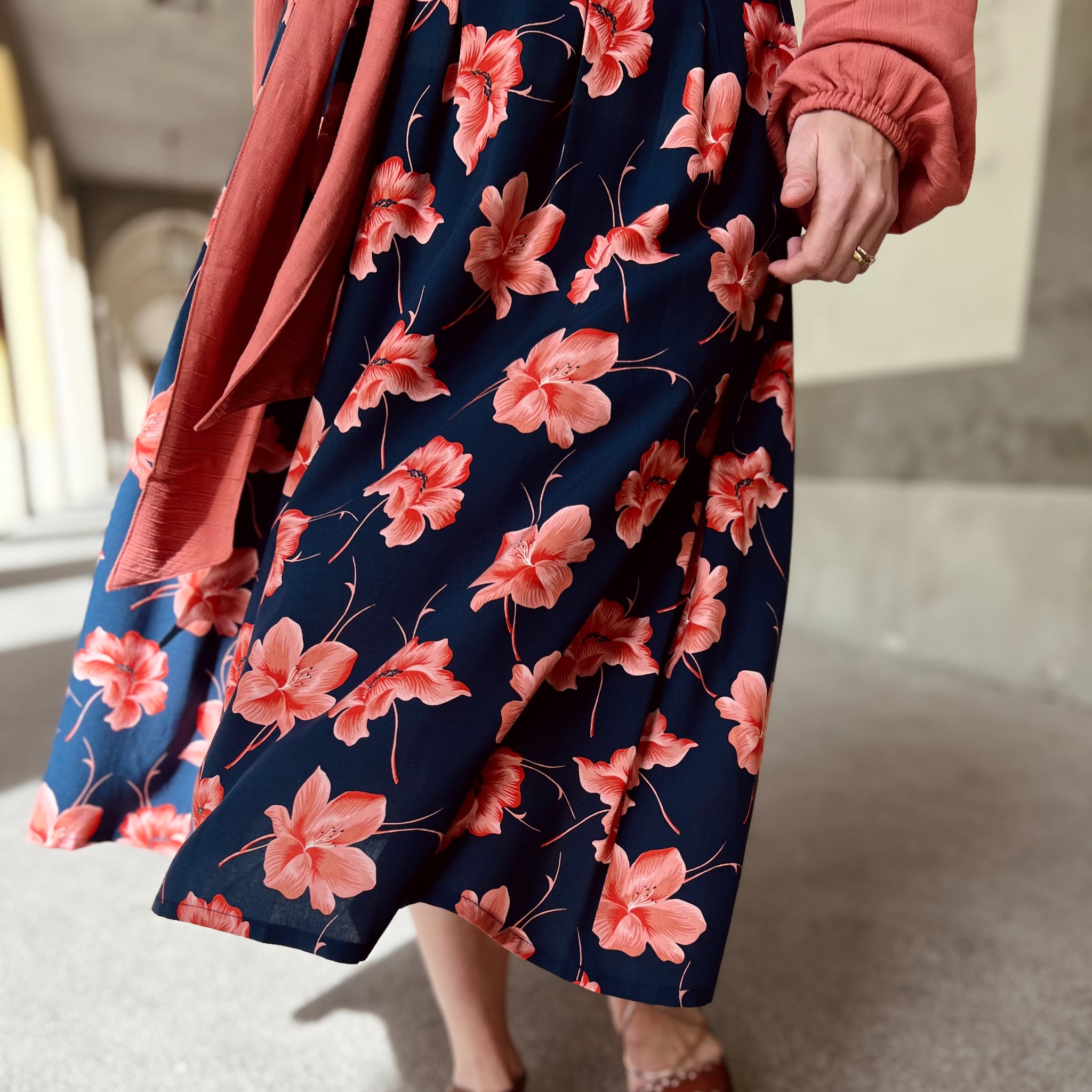 Person in a blue floral skirt with pink flowers and a coral-colored top walks on a stone pathway.