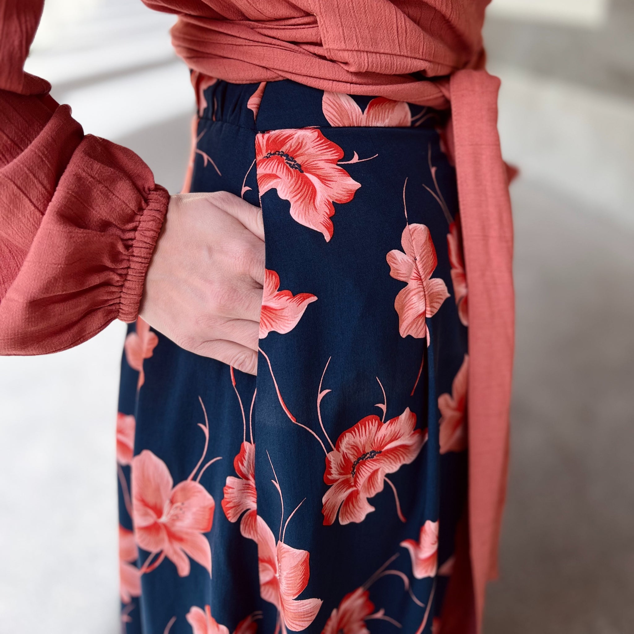 Eine Person trägt einen blau-roten Blumenrock mit der Hand in der Tasche und einem passenden roten Oberteil.