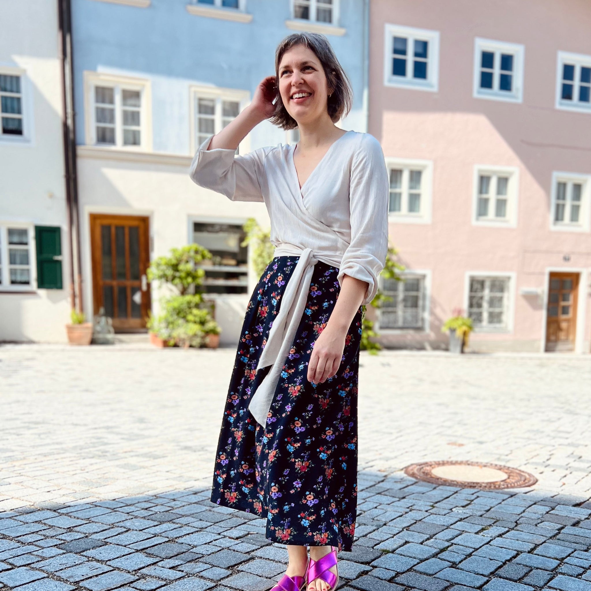 Frau in weißer Bluse und Blumenrock lächelt, während sie auf einem gepflasterten Platz vor bunten Häusern steht.