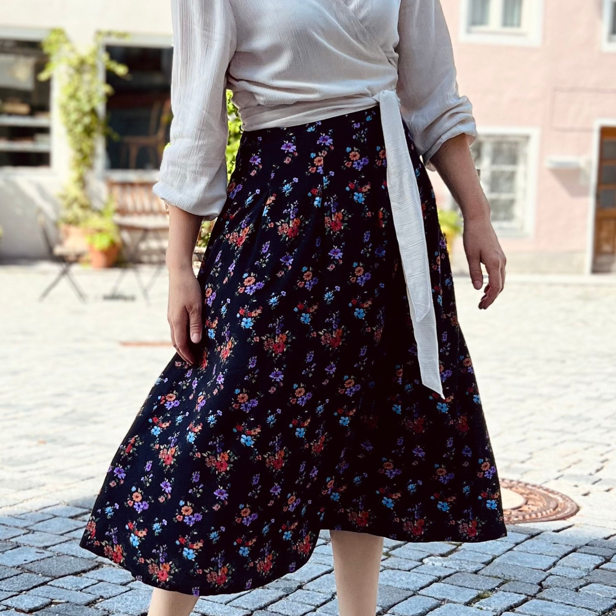 Frau in weißer Bluse und dunkelblauem Blumenkleid mit Schleife steht auf gepflastertem Platz.