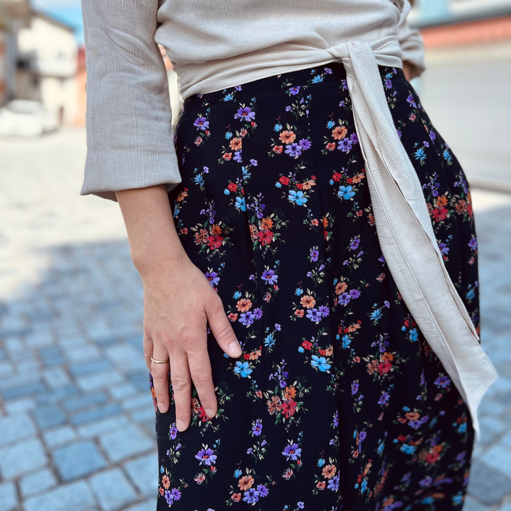 Person in beiger Bluse und schwarzem Rock mit buntem Blumenmuster steht auf einem gepflasterten Weg.