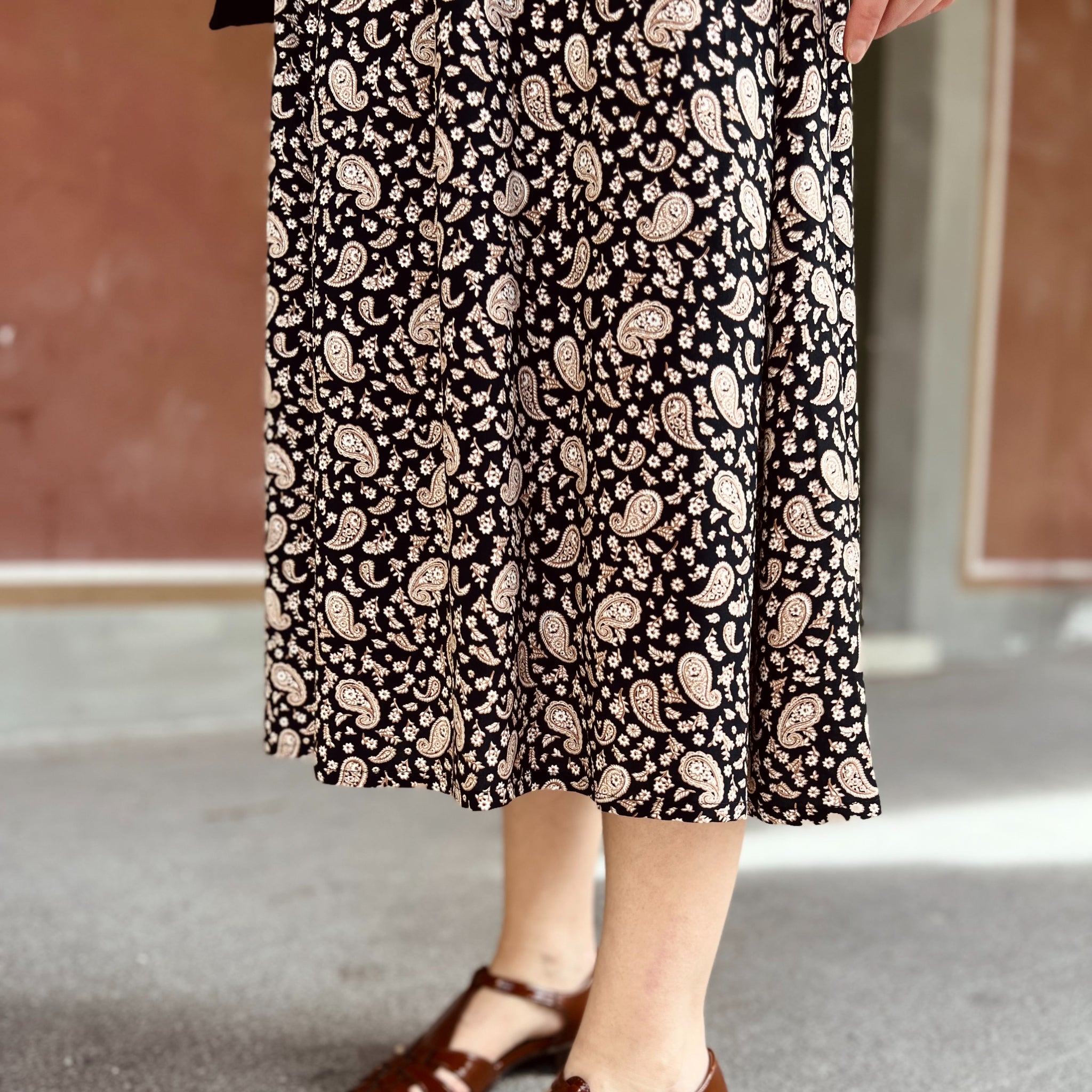Person mit einem schwarz-weiß gemusterten Paisley-Rock und braunen Sandalen steht auf grauem Boden.