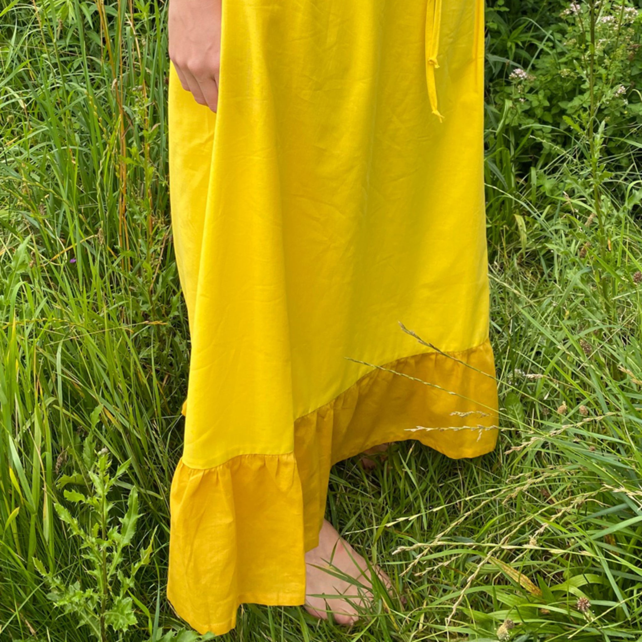 Eine Person trägt ein langes gelbes Kleid und steht barfuß im hohen Gras.