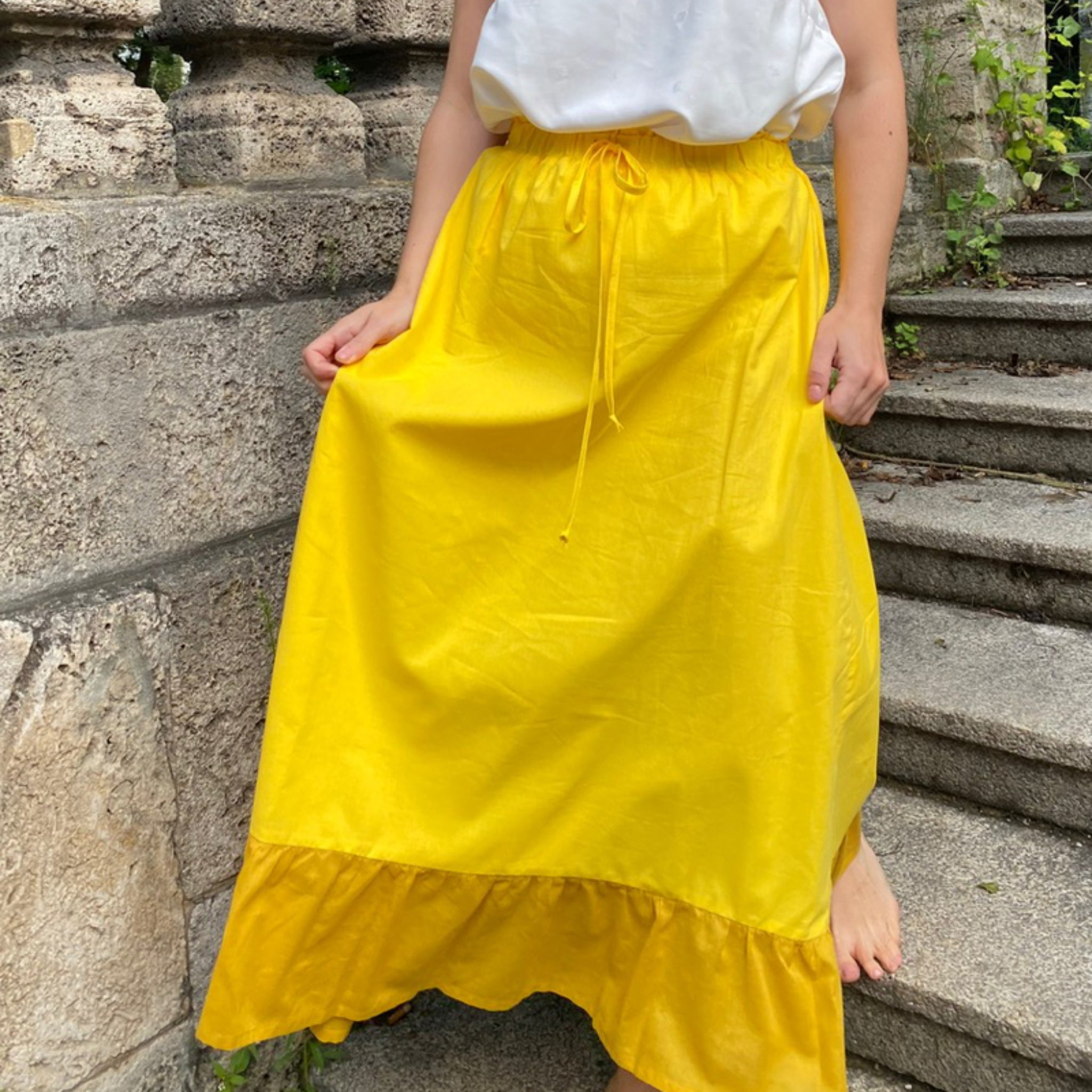 Person in a white top and bright yellow skirt stands barefoot on stone steps beside a textured wall.