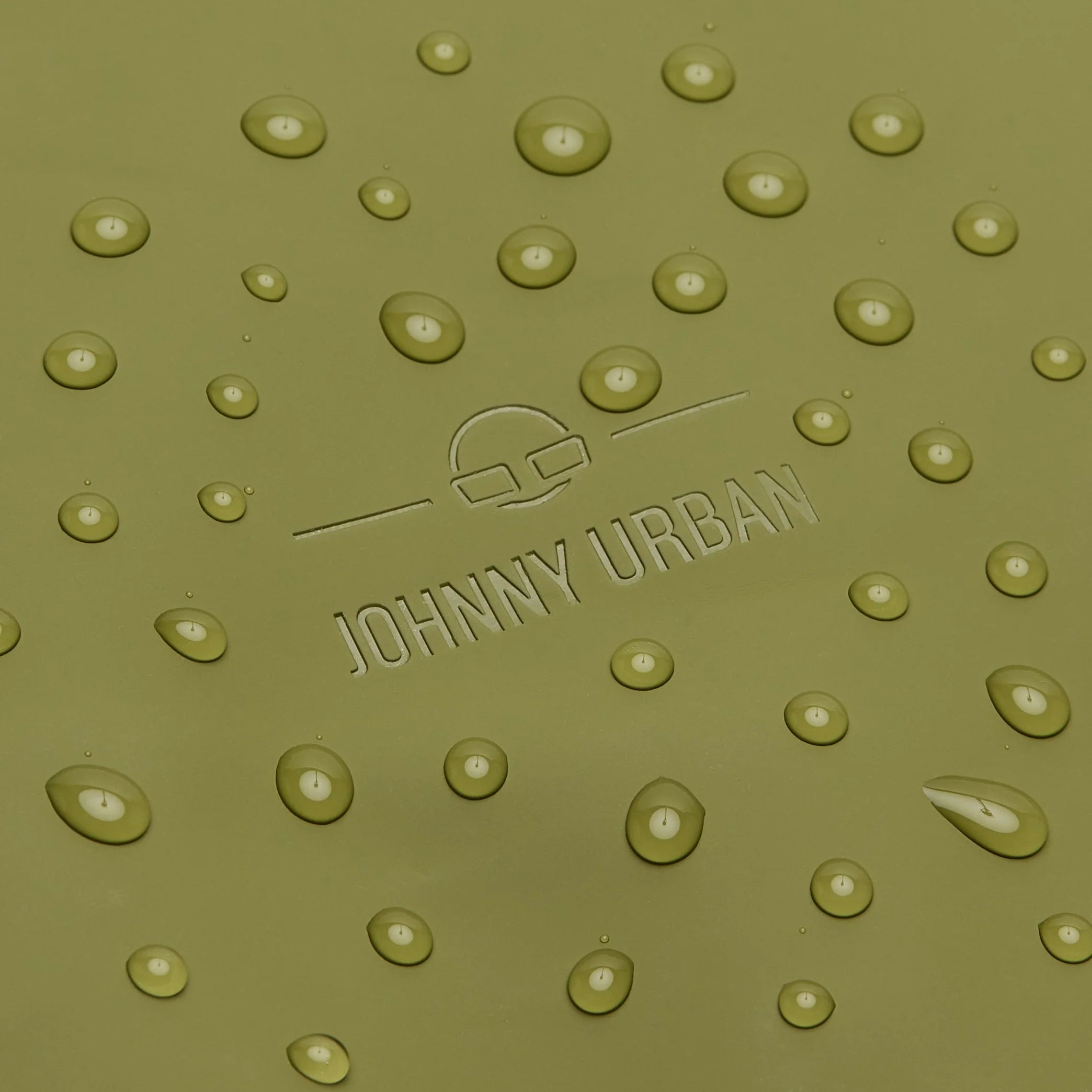 Wassertropfen auf einer glatten olivgrünen Oberfläche mit geprägtem Logo und Text JOHNNY URBAN sowie einem minimalistischen Symbol über dem Namen.