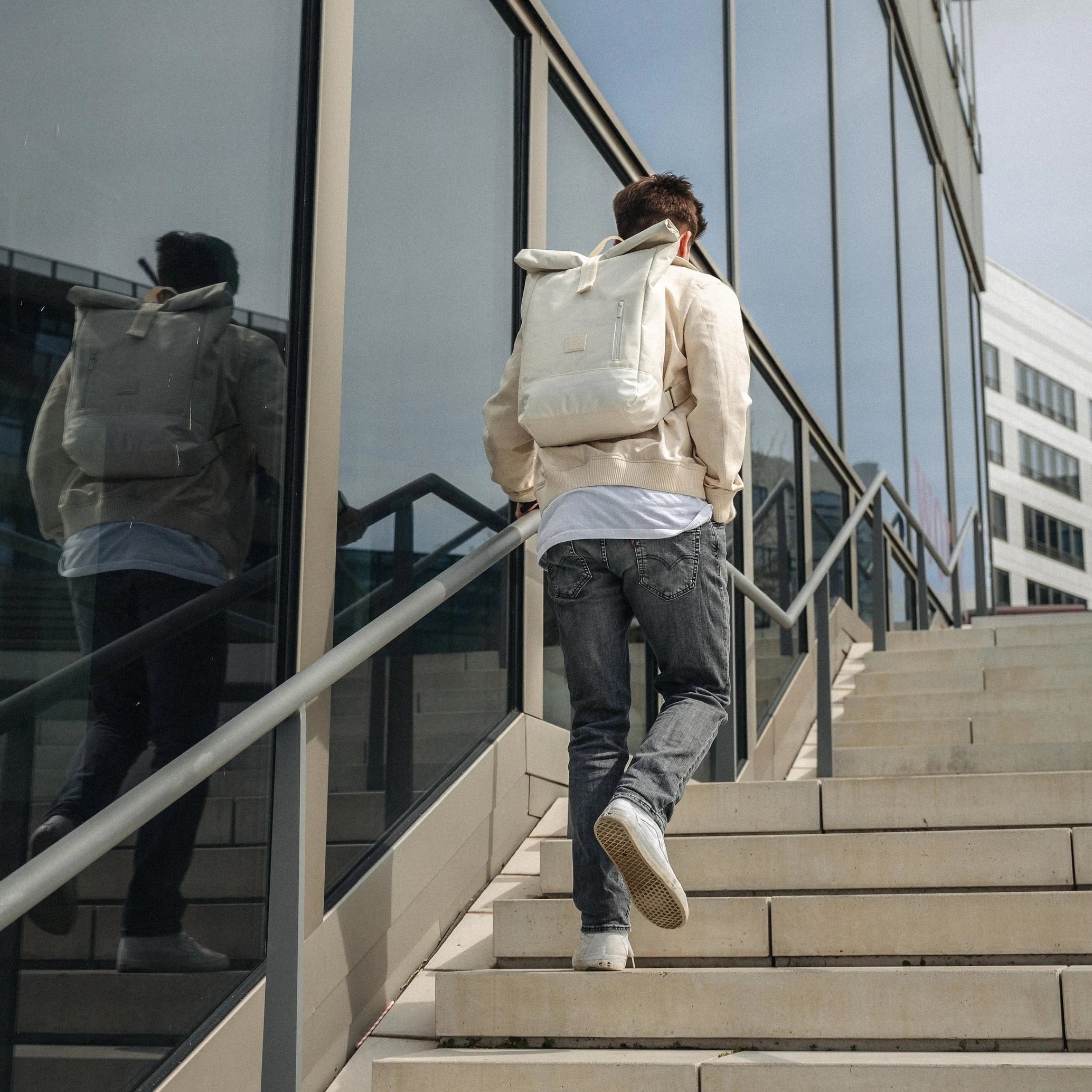 Eine Person, die eine helle Jacke, Jeans und einen weißen Rucksack trägt, geht eine Außentreppe neben einem modernen Glasgebäude hinauf.