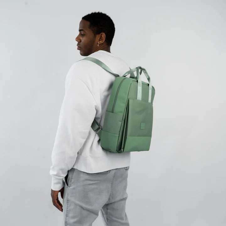 Eine Person, die ein weißes Sweatshirt und eine hellgraue Hose trägt, steht seitlich und schaut über ihre Schulter. Sie hat einen hellgrünen Rucksack auf dem Rücken. Der Hintergrund ist einfarbig und hell gefärbt.