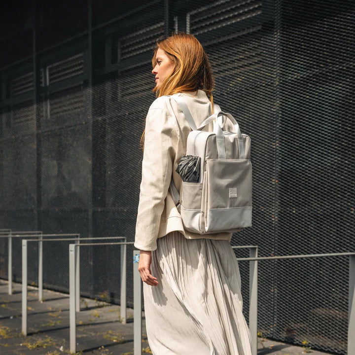 Eine Frau mit langen Haaren, heller Kleidung und Rucksack geht im Freien neben einem dunklen, modernen Gebäude mit Metallzaun und Sonnenlicht, das Schatten wirft.
