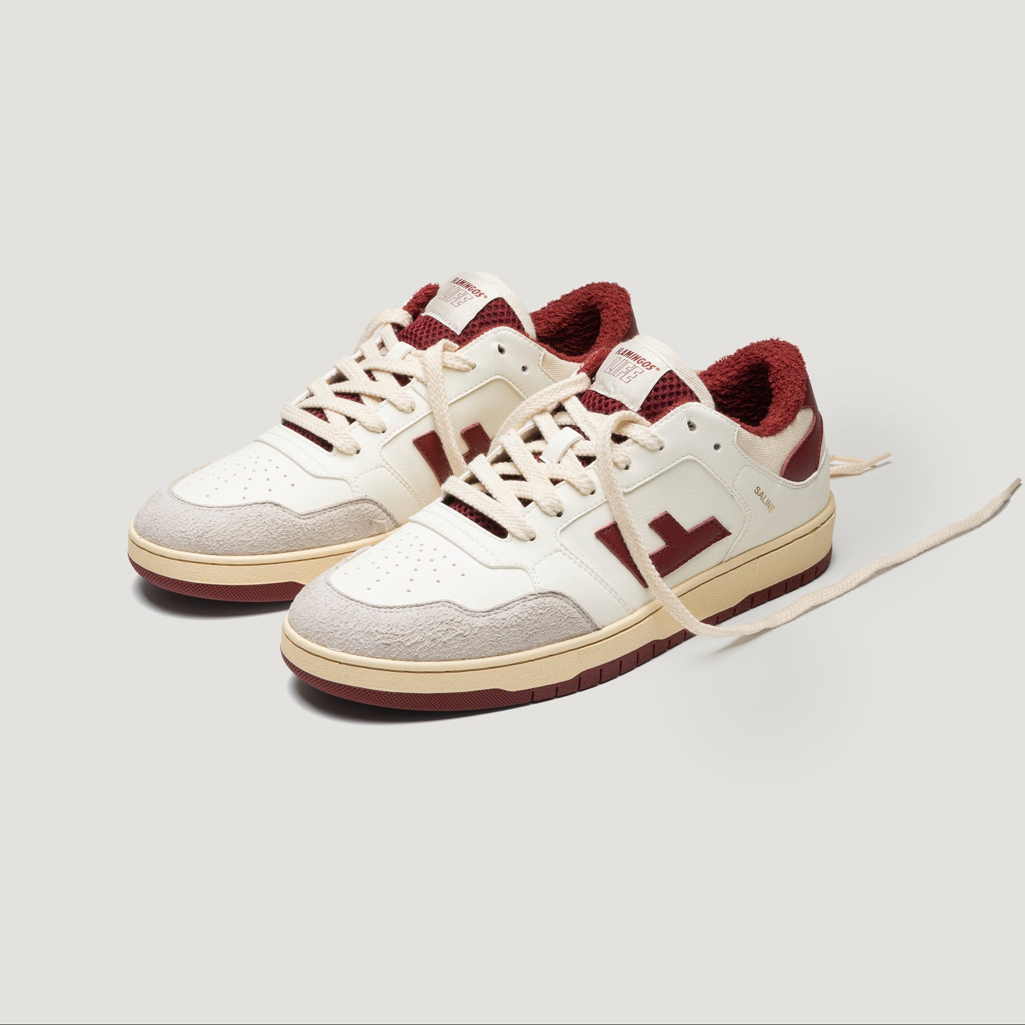 Ein Paar cremefarbene und kastanienbraune Turnschuhe mit Akzenten aus Wildleder, kastanienbraunen Details am Innenfutter und am Logo sowie beigen Gummisohlen. Die Schnürsenkel sind locker gebunden, wobei ein Schnürsenkel teilweise verlängert ist.