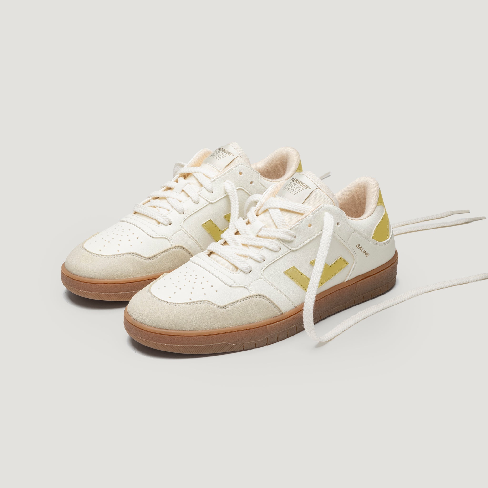 Ein Paar weiße und hellbeige Turnschuhe mit gelben Akzenten, Gummisohlen und weißen Schnürsenkeln auf einem schlichten hellen Hintergrund.