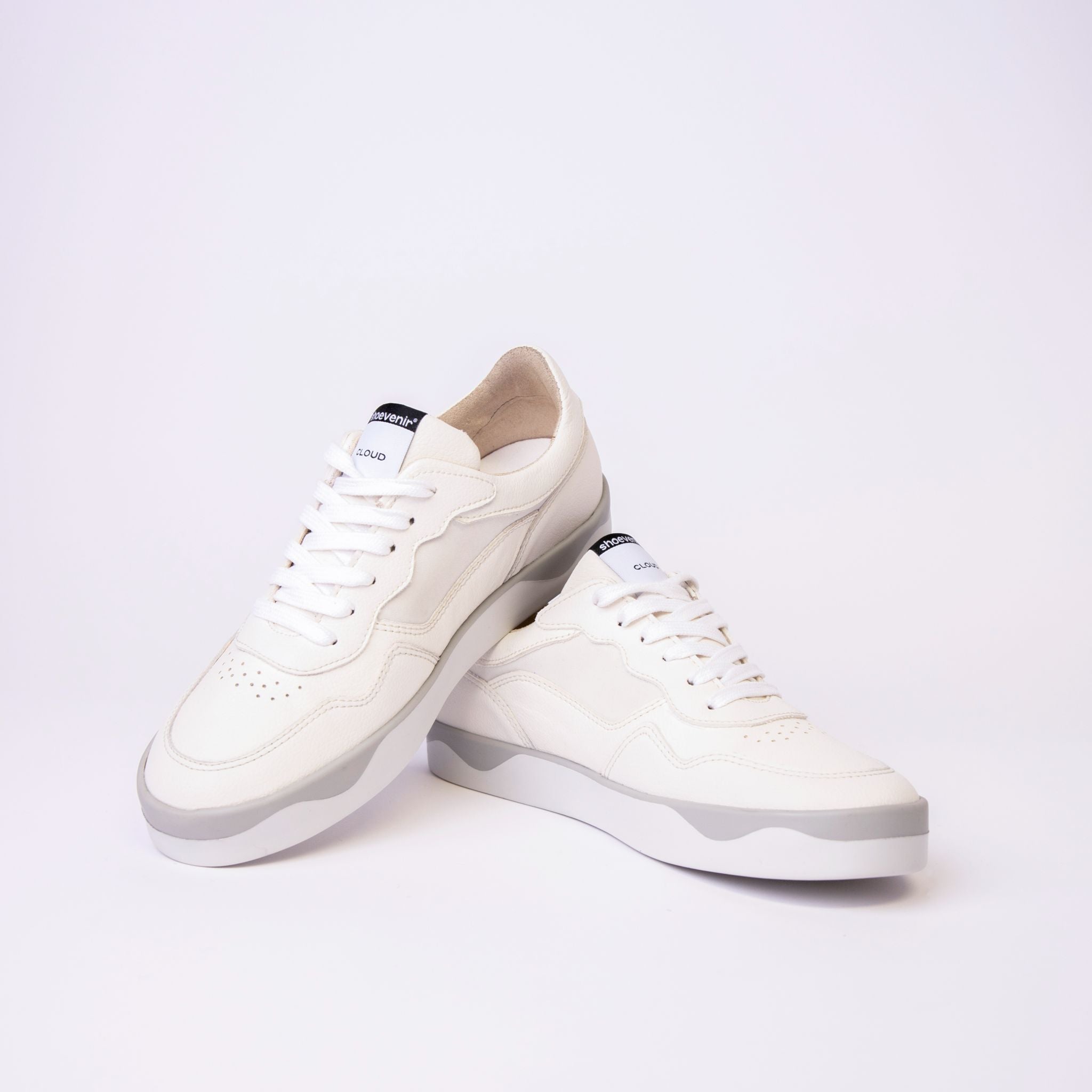 Ein Paar weißer Low-Top-Sneaker mit weißen Schnürsenkeln und hellgrauen Sohlen, die vor einem schlichten weißen Hintergrund gezeigt werden. Ein Schuh steht aufrecht, während der andere gekippt ist und auf ihm ruht.