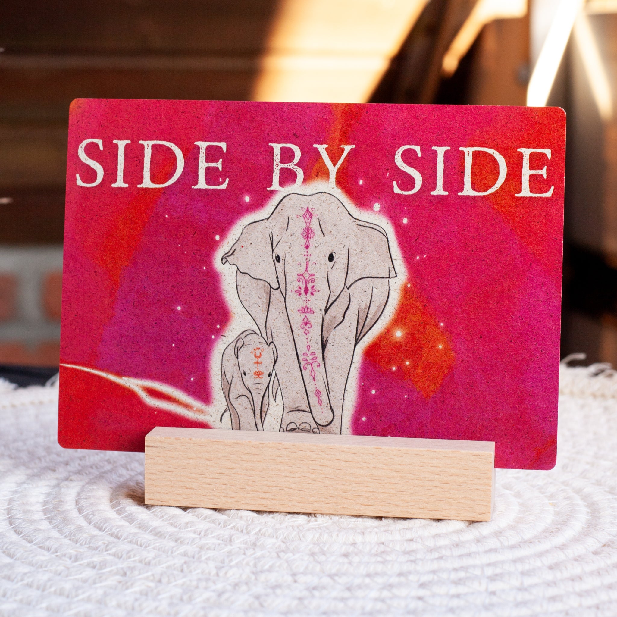 Eine Karte mit einer Illustration von zwei Elefanten, einem großen und einem kleinen, verziert mit rosa Mustern und der Aufschrift SIDE BY SIDE auf rosa-rotem Hintergrund, auf einem Holzständer.