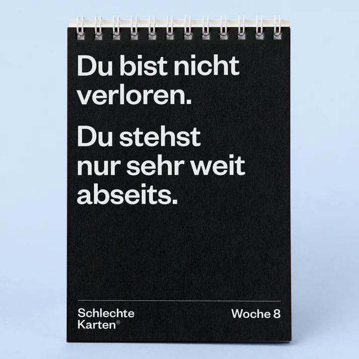 Ein schwarzer Spiral-Klappkalender zeigt einen deutschen Text: Du bist nicht verloren. Du stehst nur sehr weit abseits. Darunter steht links "Schlechte Karten®" und rechts "Woche 8" auf hellblauem Hintergrund.