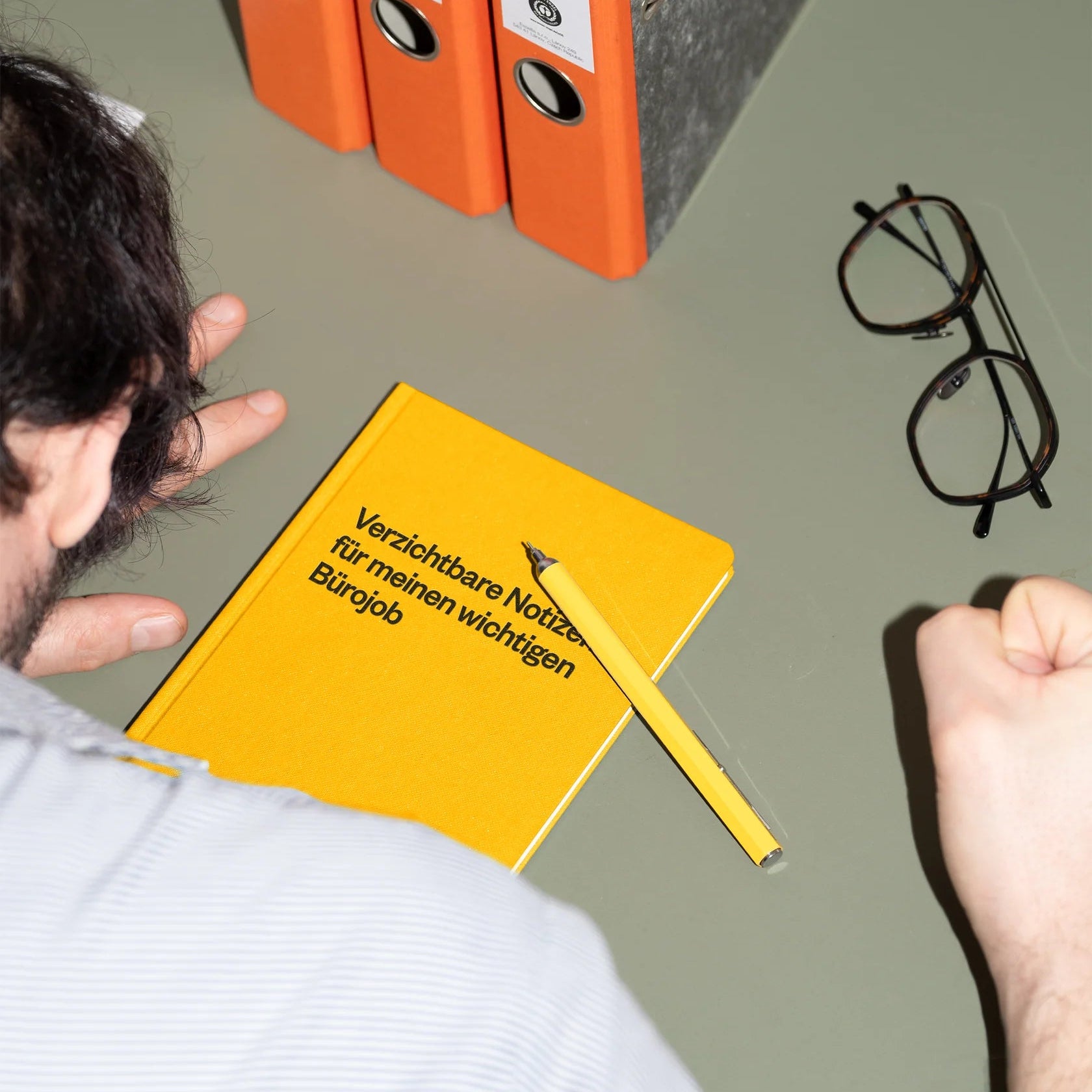 Eine Person sitzt an einem Schreibtisch mit einem gelben Notizbuch, einem gelben Stift, einer Brille und drei orangefarbenen Ordnern. Auf dem Notizbuch steht "Verzichtbare Notizen für meinen wichtigen Bürojob" auf Deutsch.