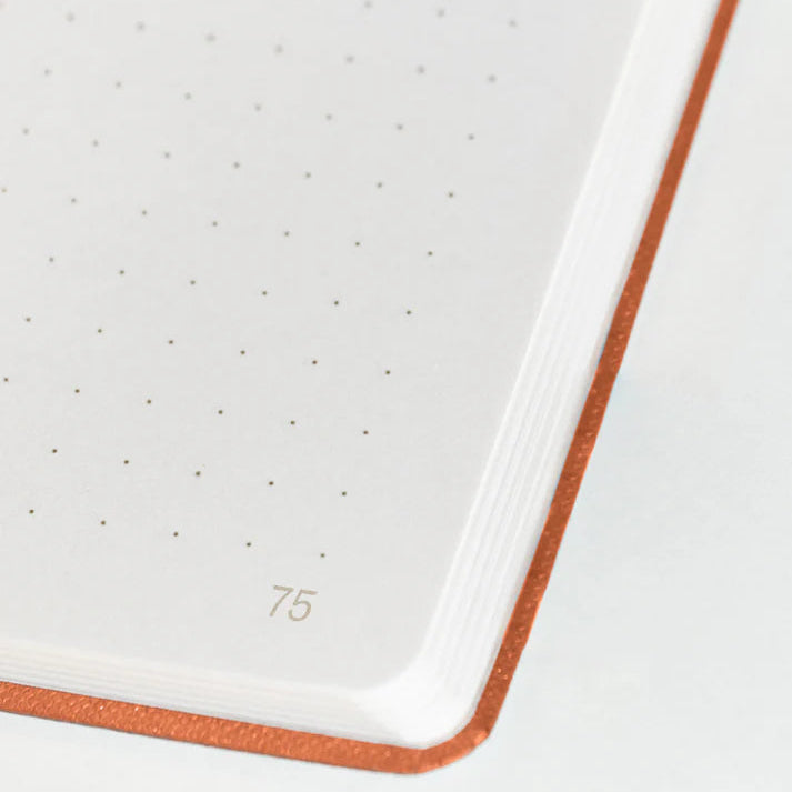 Nahaufnahme der Ecke eines aufgeschlagenen Notizbuchs mit gepunktetem Rasterpapier, das die Seitenzahl 75 und einen orange-braunen Einband zeigt.