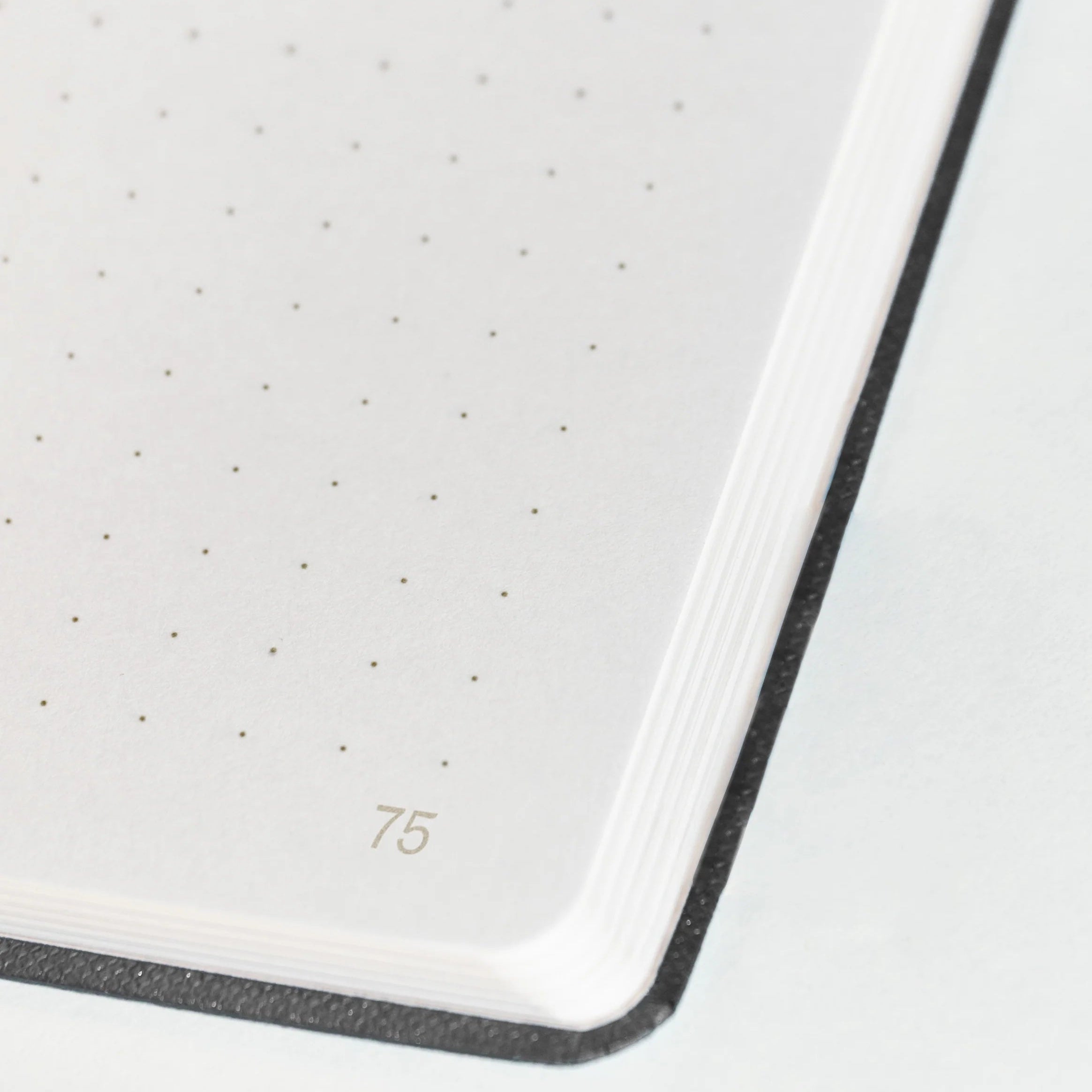 Eine Nahaufnahme der Ecke eines aufgeschlagenen Notizbuchs zeigt weißes, gepunktetes Papier mit der hellgrau gedruckten Seitenzahl 75 und einem schwarzen Umschlagrand.