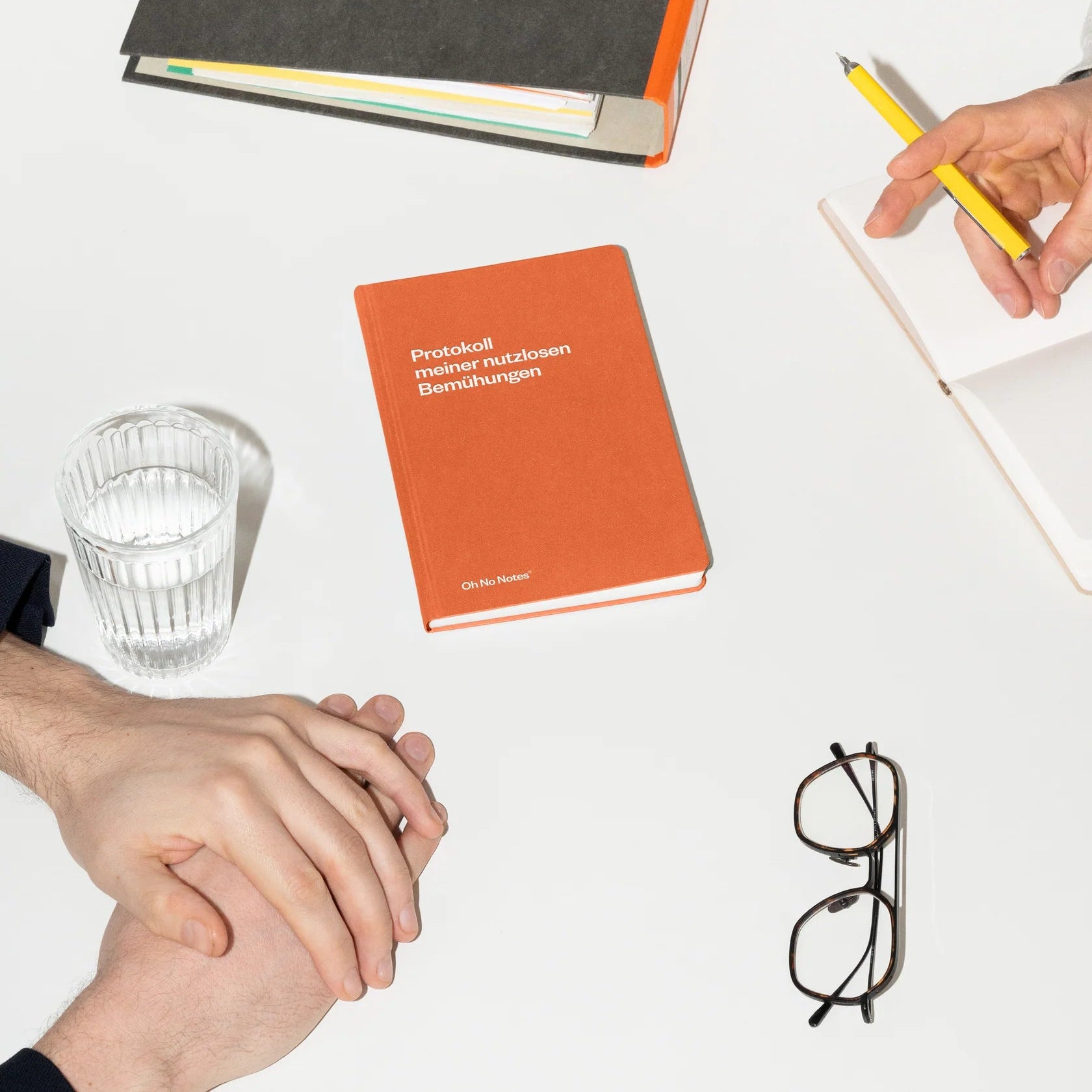 Zwei Personen sitzen an einem weißen Tisch mit einem geschlossenen roten Buch, einem Glas Wasser, einer Brille, einer schwarzen Mappe und einem Notizbuch. Eine Person schreibt mit einem gelben Bleistift, während die andere ihre Hände zusammenlegt.