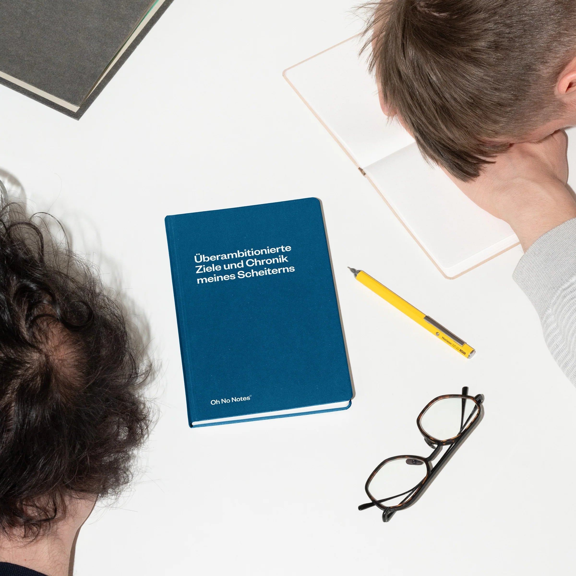 Zwei Personen sitzen an einem weißen Schreibtisch und haben ihre Köpfe frustriert gesenkt. Auf dem Schreibtisch liegen ein blaues Notizbuch, eine Brille, ein gelber Stift, ein geschlossener Laptop und eine Tastatur. Der deutsche Titel des Notizbuchs verweist auf das Scheitern.