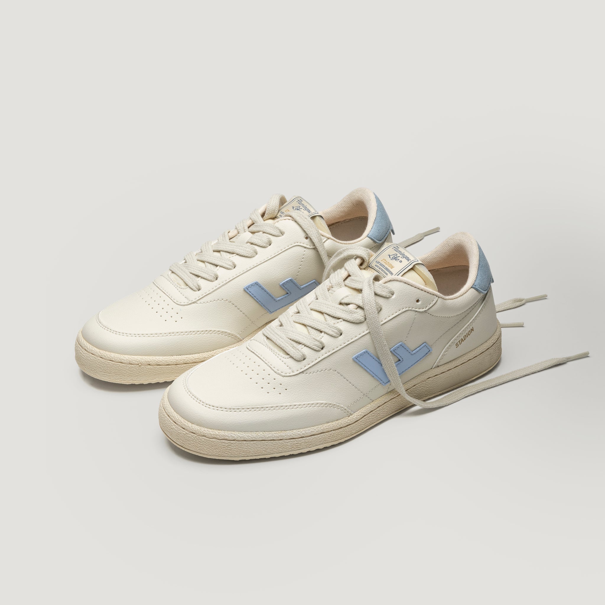 Ein Paar cremefarbene Turnschuhe mit hellblauen Akzenten, mit einem blauen L-Logo an der Seite und cremefarbenen Schnürsenkeln auf einem hellgrauen Hintergrund.