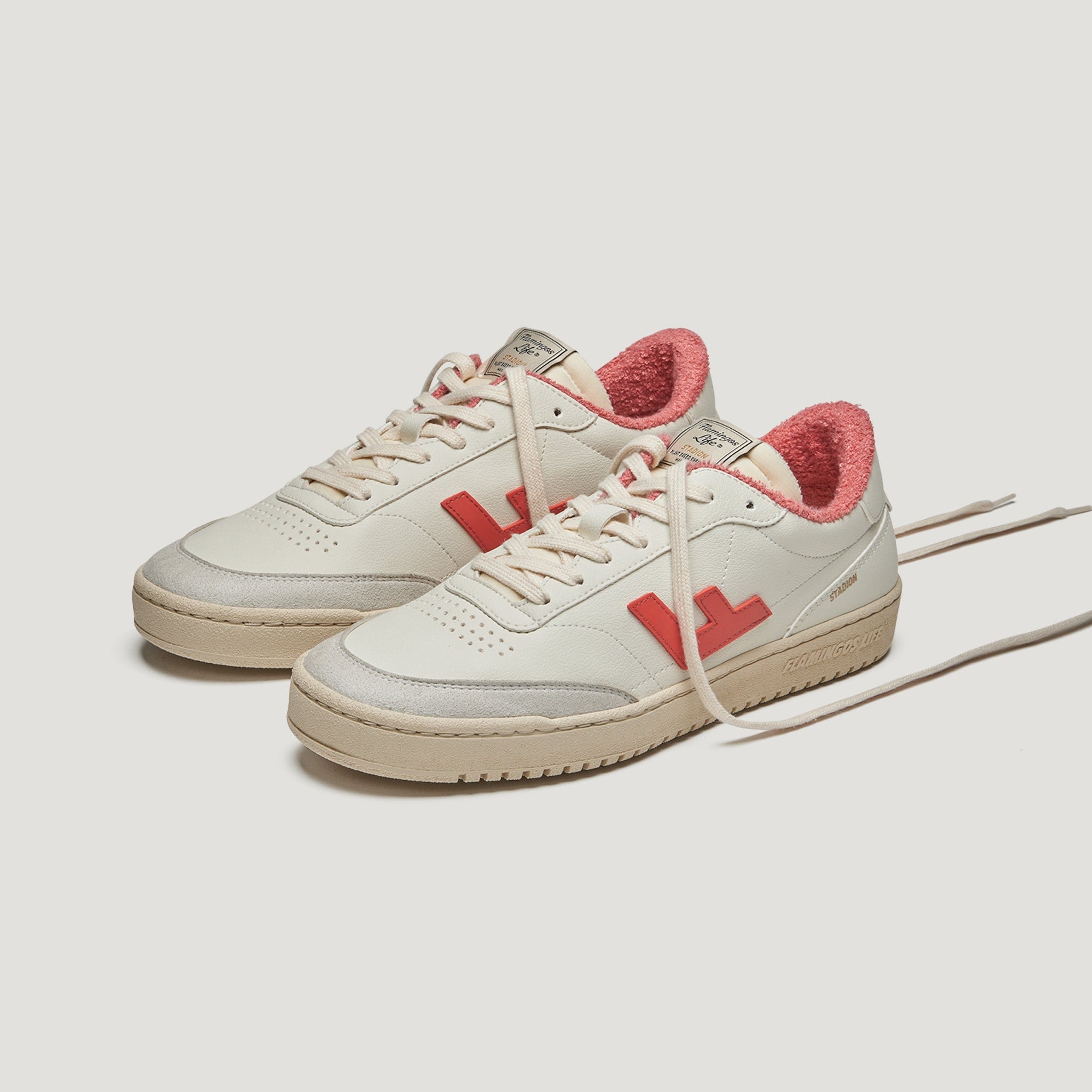 Ein Paar cremefarbene Turnschuhe mit roten Akzenten, rotem Innenfutter und beigen Sohlen. Die Schuhe haben Schnürsenkel und sind mit einem roten L-Logo an der Seite versehen. Die Schuhe sind auf einem weißen Hintergrund platziert.