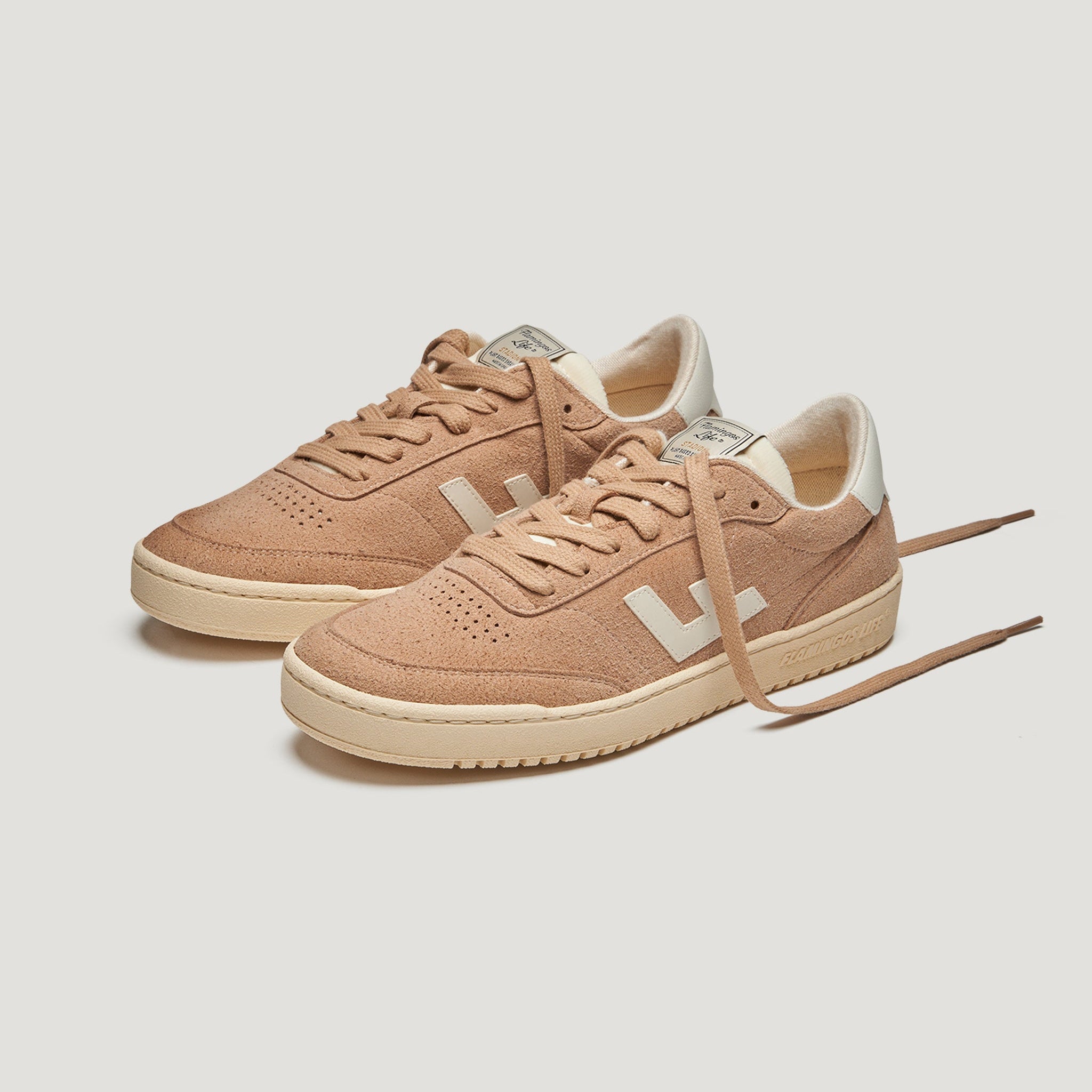 Ein Paar hellbraune Wildleder-Sneakers mit cremefarbenen Sohlen, weißen Fersenakzenten und weißen V-Logos an den Seiten. Ein Schuh hat einen offenen Schnürsenkel. Der Hintergrund ist einfarbig weiß.