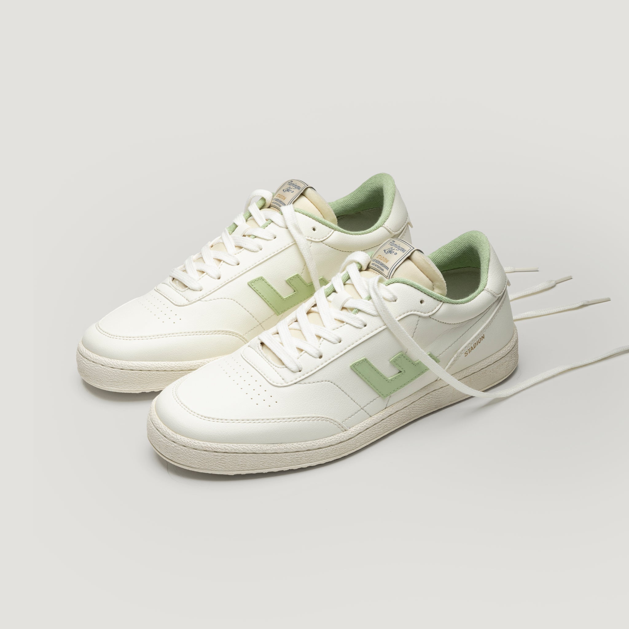 Ein Paar weiße Turnschuhe mit hellgrünen Akzenten, einschließlich des Innenfutters und eines seitlichen L-Logos, das auf einem hellgrauen Hintergrund dargestellt ist. Die Schuhe haben weiße Schnürsenkel und ein klassisches, Low-Top-Design.