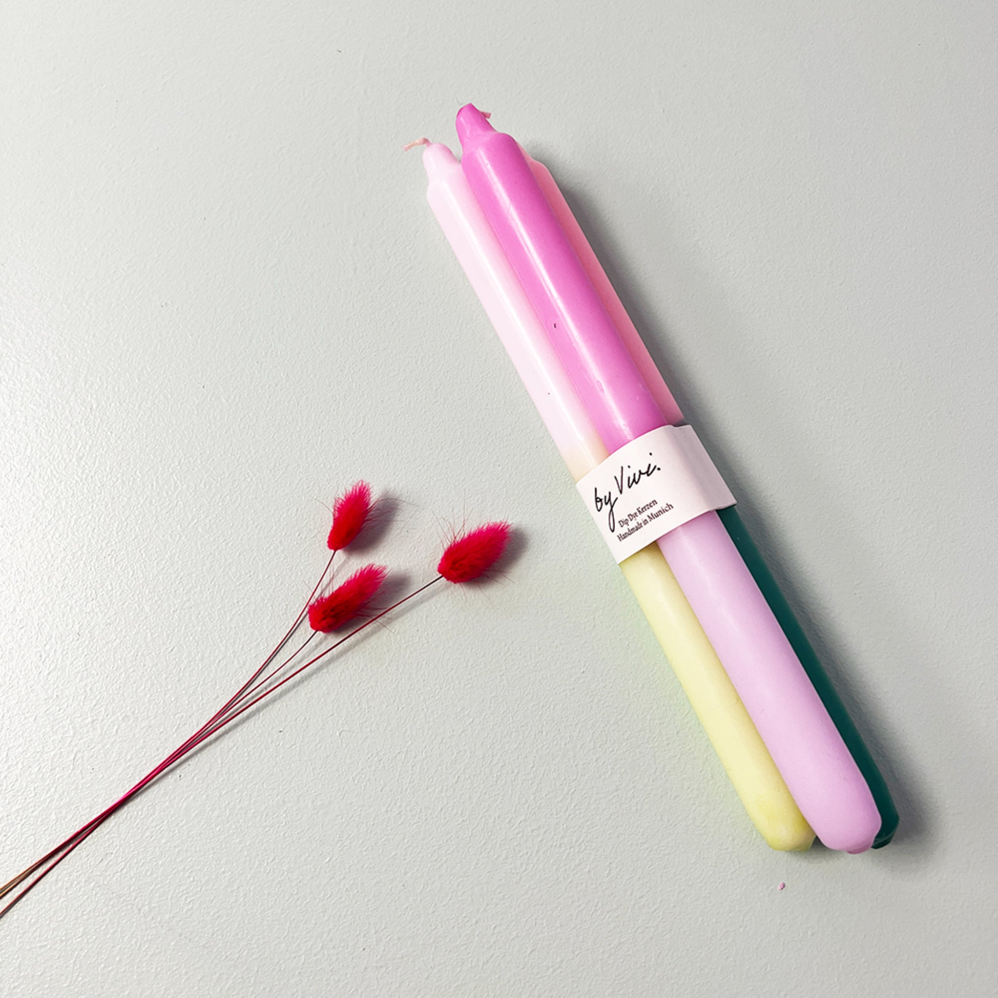 Drei pastellfarbene Kerzen liegen auf einer Oberfläche neben drei roten Trockenblumen.